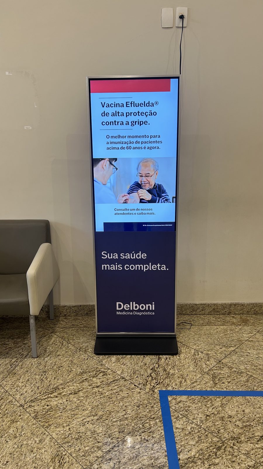 Totem digital LabDOOH em clínica Delboni exibindo campanha de vacinação contra gripe Efluelda