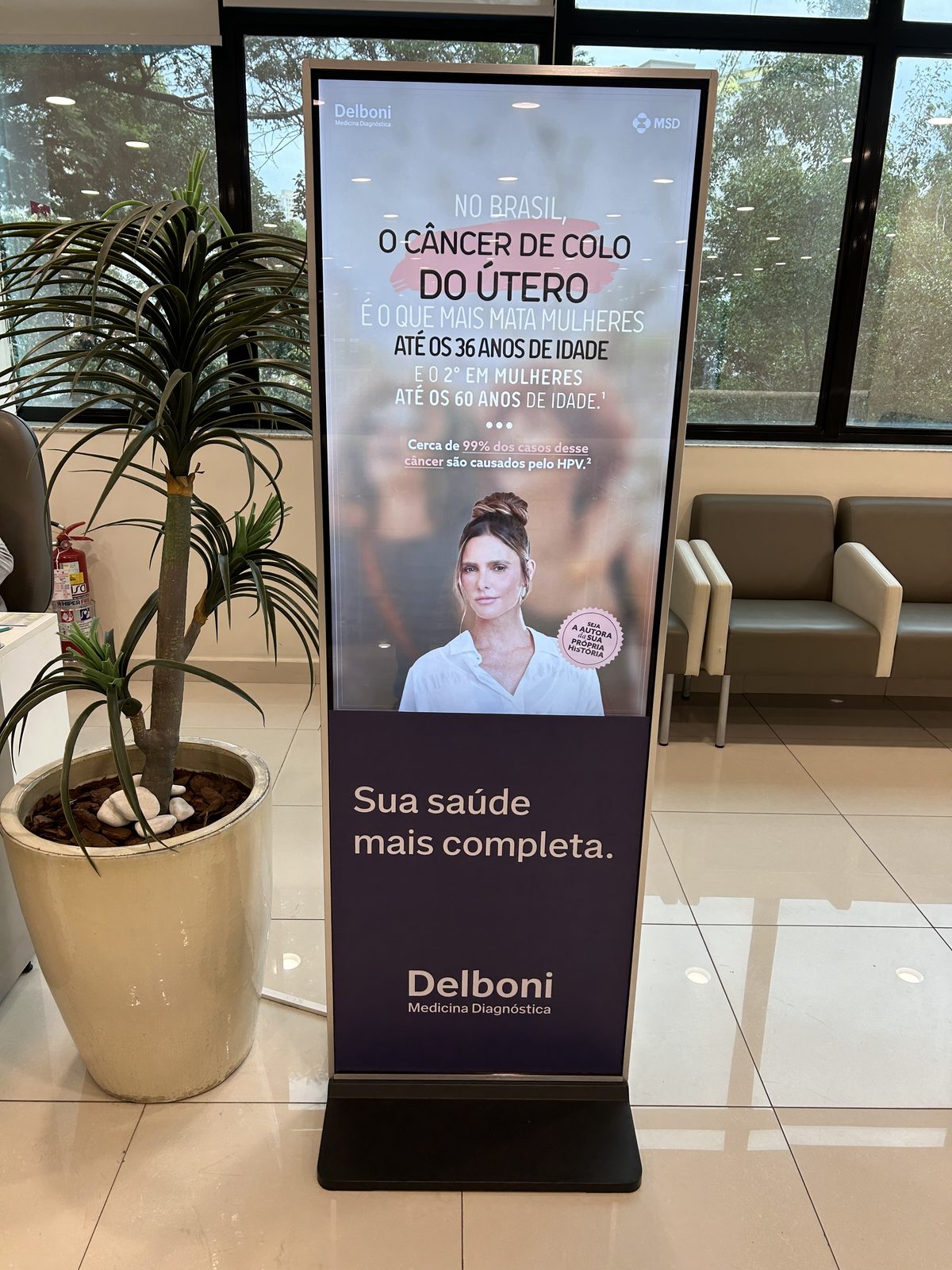 Totem LabDOOH em sala de espera com poltronas, exibindo campanha Outubro Rosa Delboni e MSD